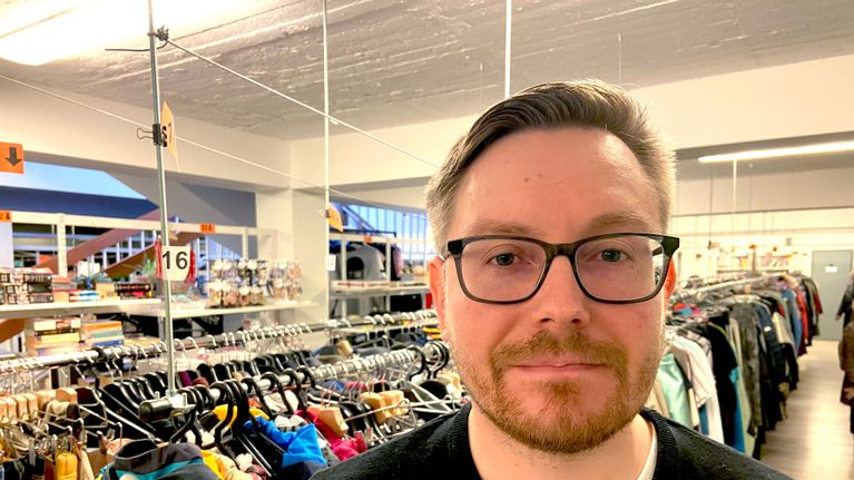 En man i glasögon, i bakgrunden kläder på hängare på ett loppis. 