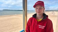 Hiekkarannalla seisoo uimavalvoja punaisessa asussa ja lippalakissa, joissa tekstit "UIMAVALVOJA" ja "LIFESAVER".