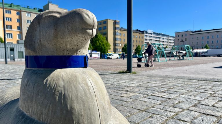 Kivinen lammas etualalla, taustalla tori, jossa kävelee ihminen rollaattorin avustamana.