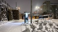 Snowy city street, a person standing at a bus stop shelter, with a pile of snow in the foreground and bus going the other direction in the background.