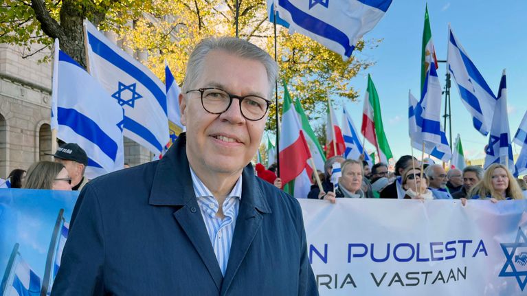Mies seisoo ulkona mielenosoituksessa Israelin lippujen ja banderollin edessä.
