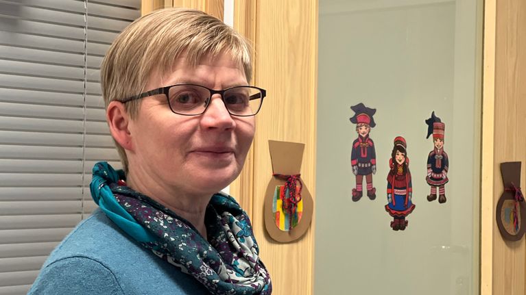 Apulaisrehtori Pirkko Olli seisoo käytävällä, taustalla saamenluokan ovi. jossa lasten tekemiä sisnapusseja. Ja kolme lapinpukuista kuvaa liimattuna luokkahuoneen oven ikkunaan, 9.1.2024 Ivalon koulussa.