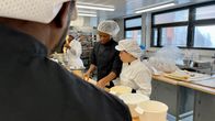 Leipurin suoja-asuun pukeutunut tummaihoinen opiskelija ja aasialaistaustainen opiskelija katsovat leivontapöytää vieressään useita taikinakulhoja.