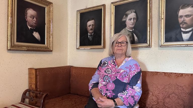 En äldre kvinna sitter på en soffa framför fyra inramade porträtt på en beige vägg.