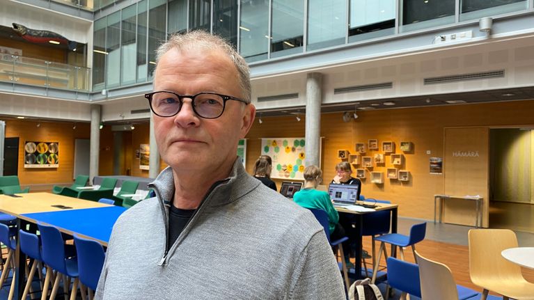 Mies katsoo kameraan. Taustalla opiskelijoita tietokoneen äärellä.