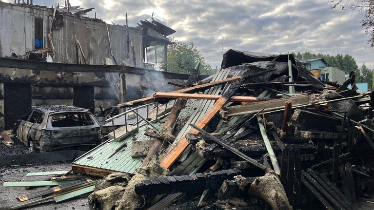 Bränt och förstört biltak, bråte från biltaket i en hög, en bränd bil och i bakgrunden en förstörd gavel av ett radhus. 