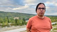 Mies seisoo ulkona mäkisellä näköalapaikalla kesällä. Taaempana näkyy tie sekä maisemassa metsää, peltoja ja kaukainen järvi.