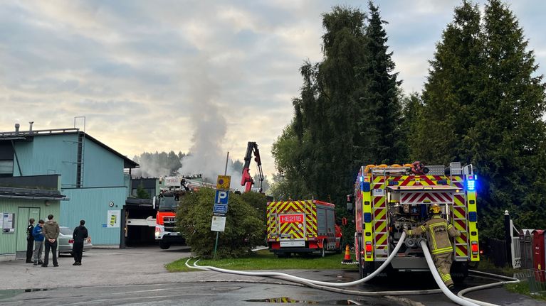 Räddningsfordon med slangar och brandmän står utanför en byggnad med rök i bakgrunden.