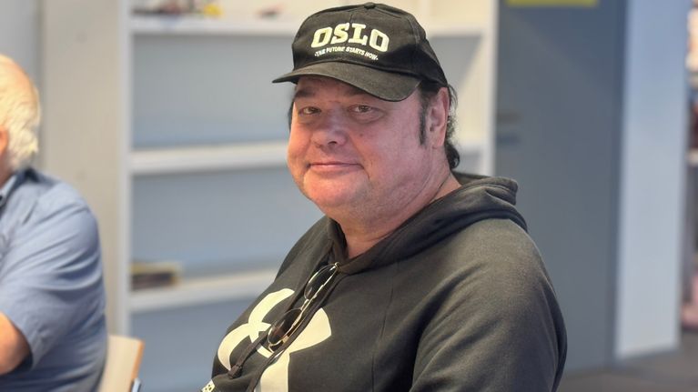 A man wearing a black "OSLO" cap and a black hoodie with "ARMOUR" text sits indoors near bookshelves.