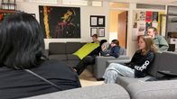 Mustatukkainen ihminen istuu harmaalla sohvalla selin, keskellä ja kuvan oikeassa reunassa on kolme nuorta, jotka katsovat televisiota, yksi heistä levittää vihreää fleece-vilttiä polvilleen. Kuvan oikeassa reunassa vihreään paitaan pukeutunut parrakas ja pitkätukkainen mies seuraa tilannetta hymyillen.