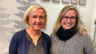 Lähikuvassa kaksi vaaleahkohiuksista naista, vasemmalla polkkatukkainen nainen sinisessä villatakissa ja oikealla hiukan nuorempi silmälasipäinen ja pitkätukkainen nainen. Molemmat hymyilevät. 