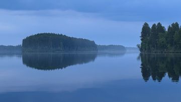 Tyyni järvi, jonka rannalla on metsää ja taustalla puustoinen saari, sinertävän sumun keskellä.