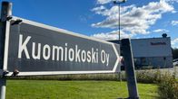 Kuomiokoski-yrityksen kyltti, taustalla näkyy tehdas, jossa lukee Kuoma.
