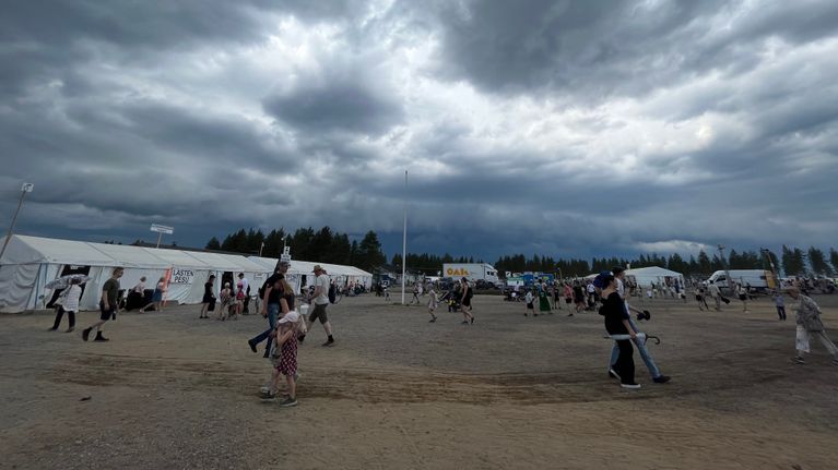 Synkät myrskypilvet nousevat horisontissa hiekkakentän yllä. Ihmiset kävelevät ulkona.