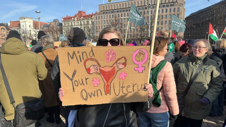 En person håller upp en skylt med texten "Mind Your Own Uterus" tillsammans med en illustration av en livmoder och venussymboler.