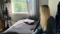 Nuori nainen istuu sängyllä ja katsoo ikkunasta ulos. Ulkona näkyy vihreitä puita. Naisen kasvoja ei näy.