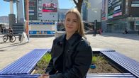 Mustaan nahkatakkiin pukeutunut nuori nainen istuu sinisellä penkillä. Hänen takanaan näkyy kaupungin kävelykatua.