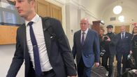 Useita pukuihin pukeutuneita henkilöitä kävelee käytävällä, taustalla kameramiehiä ja muita ihmisiä.