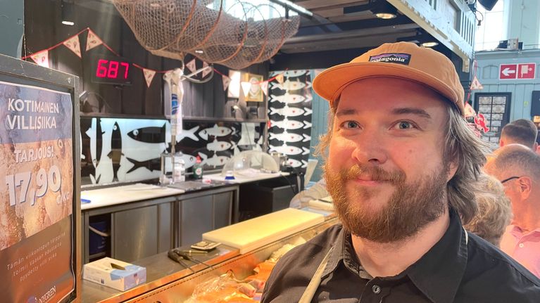 Parrakas ja aavistuksen pitkähiuksinen mies oranssi lippis päässä katsoo kameraan hymyillen. Taustalla näkyy Tamperen kauppahallin asiakkaita ja kalatiski.