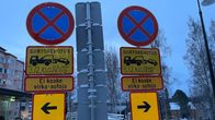 Kaksi pysähtymiskieltomerkkiä, joiden alapuolella on ajoneuvon siirtokehoitus-merkki. Taustalla luminen maisema ja taloja.