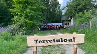 Tervetuloa-kyltin takana näkyy maalaistalon pihapiiri, missä on paljon ihmisiä.