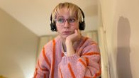 En person sitter inomhus iklädd en rosa och orange randig tröja och svarta hörlurar.