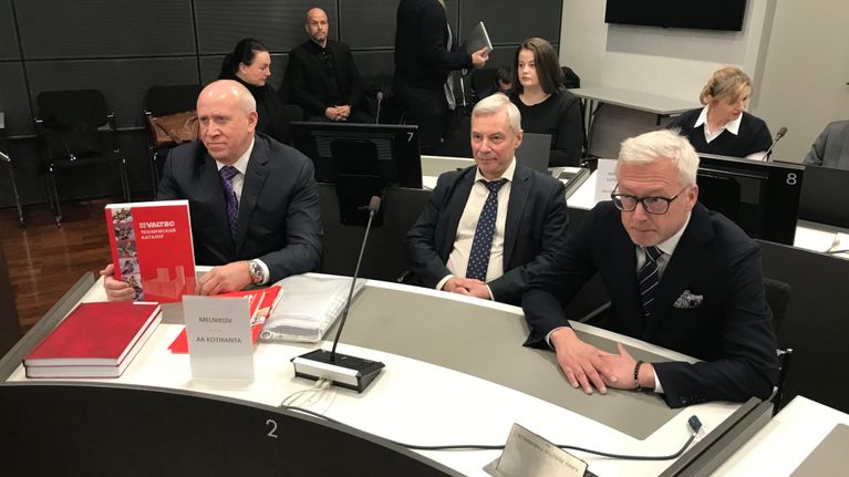 I en rättssal sitter tre män i mörka kostymer bakom ett bord.