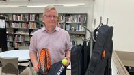 Vaalea silmälasipäinen mies seisoo kirjastossa ja pitelee kädessään lainattavaa padel-mailaa ja palloa.