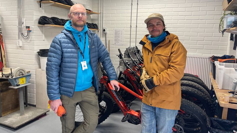 Two men wearing outdoor clothing lean against a row of fat-tyre bikes in a storeroom, with a row of helmets behind.