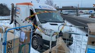 Pakettiauto, jossa lukee "Enset Oy", on ajautunut lumen peittämälle alueelle. Tien vieressä on pahvilaatikoita ja metalliaidat, jotka on ympäröity varoitusnauhalla. Taustalla näkyy maantie ja siltoja.