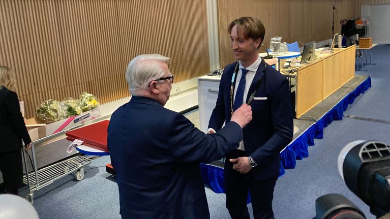Harmaahiuksinen mies kättelee nuorta miestä valtuustosalin edessä kameroiden kuvatessa heitä.