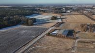 Kuvassa on talvinen maalaismaisema, jossa näkyy peltoja, metsää ja rakennuksia. Maassa on ohut lumikerros ja peltojen väleissä kulkevat tiet risteävät etualalla. Horisontissa erottuu metsää ja kauempana näkyy teollisuusrakennuksia tai muita suurempia rakennuksia.