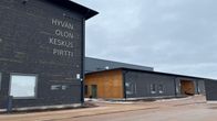 Pudasjärven Hyvän olon keskus Pirtin tummanharmaata ja keltaista hirsijulkisivua, taustalla näkyy harmaa pilvinen kevättalven taivas.