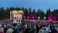Pori jazzin banderolli keskellä ihmispaljoutta Porin kirjurinluodon areenalla hämärtyvässä illassa taustalla kuuset valaistu pinkeiksi.