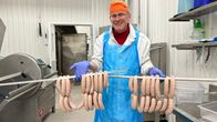 Valkoiseen takkiin ja siniseen muoviessuun pukeutunut silmälasipäinen mies pitelee käsissään kahta metallitankoa, joissa roikkuu kymmeniä vaaleita makkarapötköjä. Taustalla teräksenharmaita hyllyjä, ovia ja laitteita.