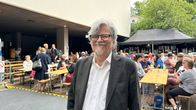 M. A. Numminen mustassa takissaan. Taustalla ihmisiä ruokapöydissä. 