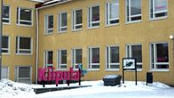 Lähikuvassa on keltainen betonitalo, jonka pihassa on punainen logoteksti.