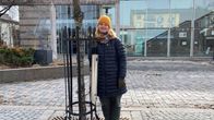 Nainen seisoo nojaten kävelykadun puun kehikkoon. Kehikossa on kiinnitettynä pystymallinen teräksinen tumppiroskis.
