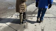 Kaksi ihmistä kävelee asfalttikatua. Vasemman puoleisella beiget vaatteet ja kävelysauvat, oikean puoleisella siniset vaatteet.