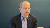 Kuvassa on henkilö pukeutuneena tummansävyiseen pukuun ja vaaleansävyiseen kauluspaitaan. Tausta on yksivärinen ja sinertävä.