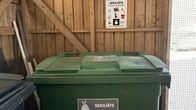 Kuvassa on ruskeaseinäinen jätekatoksen sisätiloihin sijoitettu vihreä seka-astian jäteastia, jossa symbolikuvio ja teksti "SEKAJÄTE"
