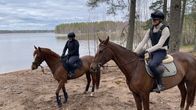 Kuvassa kaksi ratsastajaa hevostensa selässä järven rannalla metsäisessä maisemassa. 