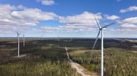 Tuulivoimalat kohoavat metsän yläpuolella aurinkoisena päivänä, taustalla on sininen taivas ja kumpupilviä.