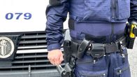 Lähikuvassa on poliisin virka-asu, jossa näkyvät varusteet. Seisovan poliisin takana on auton keula.