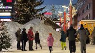 Lapset leikkivät suurella lumikasalla kaupungin valojen loisteessa, ja aikuiset seuraavat vierestä talvisissa asuissaan.