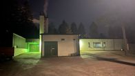 Matala vaalea rakennus iltahämärässä ulkovalot päällä. Isosta piipusta nousee savua. Taustalla mustia puita.
