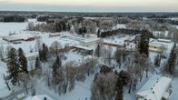Talvinen maisema, jossa näkyy punatiilisiä rakennuksia lumipeitteisen metsän ja peltomaiseman keskellä.