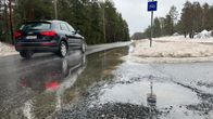 Katu, jonka toiseen reunaan on valunut runsaasti vettä lätäkköön. Tiellä ajaa musta auto. Tien reunassa vähän lunta.