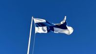 Valkoinen lippu, jossa on sininen risti, liehuu tuulessa lipputangossa. Suomen lippu on tangossa. 