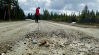 Henkilö kävelee hiekkatiellä keväisessä metsämaisemassa, tien pinnalla näkyy ruosteinen pultti.